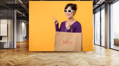 A cheerful female consumer, wearing a purple shirt and holding a shopping bag, smiles against a yellow background, portraying the happiness of shopping and making great purchases. Wall mural