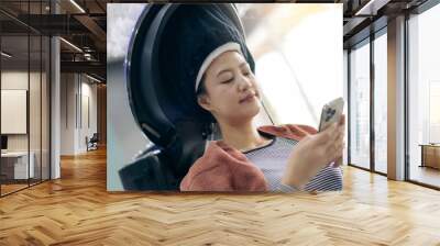 A woman relaxing in salon while using her smartphone, enjoying hair treatment. She appears calm and content. Wall mural