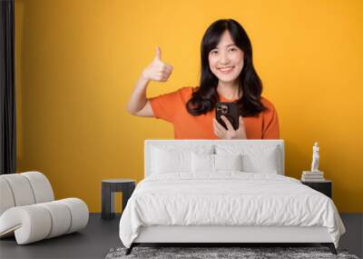 Embody joy with a young Asian woman in her 30s, donning an orange shirt, showing thumb up gesture while using smartphone on yellow background. Wall mural