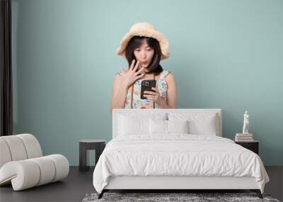 Happy Asian woman wearing casual dress and hat with camera while getting amazing news on mobile phone on vacation or travel theme isolated on pastel green background. Travel and vacation concept. Wall mural