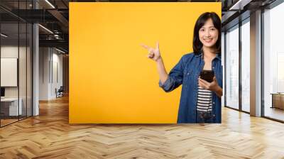 Portrait beautiful young asian woman happy smile dressed in denim jacket showing smartphone with pointing finger hand gesture to free space isolated on yellow studio background. app smartphone concept Wall mural