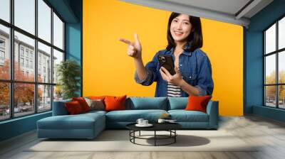 Portrait beautiful young asian woman happy smile dressed in denim jacket showing smartphone with pointing finger hand gesture to free space isolated on yellow studio background. app smartphone concept Wall mural