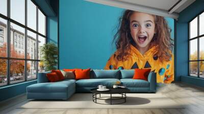 Cheerful and amazed preteen child dressed in a whimsical and festive Halloween costume standing against a plain azure blue studio backdrop with copy space above Wall mural