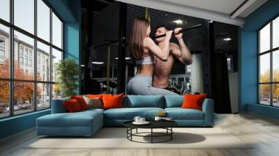 Young couple is working out at gym. Attractive woman and handsome muscular man are training in light modern gym. Shirtless man is doing pull up with woman hanging on him. Wall mural