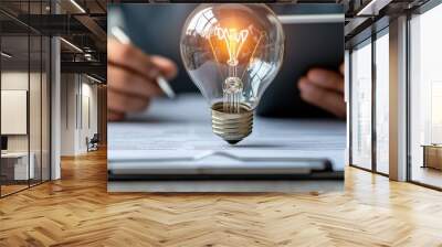 Person writing notes with a tablet and a glowing light bulb on paper, representing innovative ideas and brainstorming. Wall mural