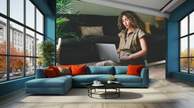 Beautiful female in casual outfit chilling on a floor beside comfortable couch in a living room with laptop on her knees. Web designer browsing  laptop while working on a remote project at home. Wall mural