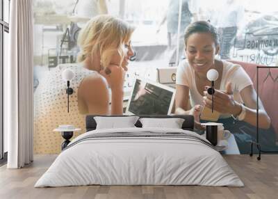 two female hipsters are watching photos on a screen of a smartphone while having rest in a coffee sh Wall mural