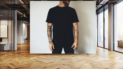 young attractive bearded man with tattoos, dressed in a black blank t-shirt, posing on a white wall background. Empty space for you logo or design. Wall mural