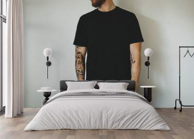 young attractive man with a beard and tattoos, dressed in a black blank t-shirt, posing against a white wall. Empty space for you logo or design. Wall mural