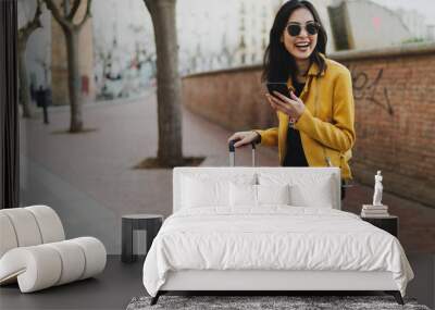 Young smiling woman traveler enjoying her first minutes in a new city and sharing the emotions with a friends online by a smartphone. Asian hipster girl reading messages on a mobile phone. Wall mural