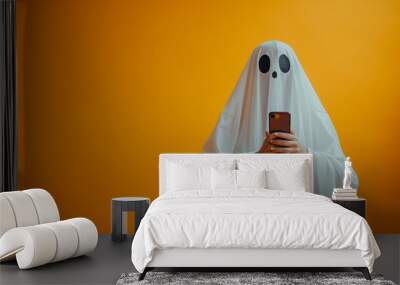 Ghost costumed person with white sheet holding smartphone on orange background. Halloween vacation concept, online sales, web, advertising and sales. Copy space. Wall mural