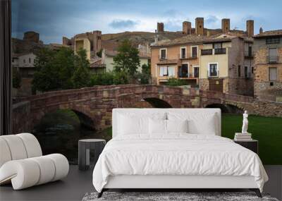 Scenic view of a medieval bridge made with reddish stone blocks and the castle on the top of the hill behind the houses on a cloudy day, Molina de Aragón, Guadalajara, Spain Wall mural