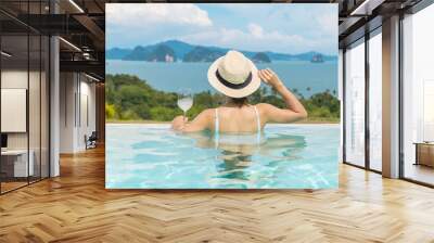 Happy woman in white swimsuit drinking wine in luxury swimming pool hotel against beautiful view, young female with hat enjoy in tropical resort. Relaxing, summer travel, holiday and vacation concept Wall mural