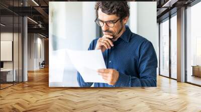 Business man wearing glasses  holding paper document. Successful male portrait thinking and reading contract at the office by the window Wall mural