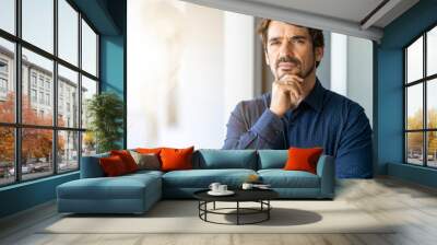 Man with hand on his chin looking at the camera. Powerful successful male portrait at the office by the window with a serious thinking expression face Wall mural