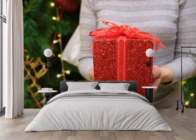 Female handing a red glitter square shape gift box wrapped with red ribbon bow to someone, blurred Christmas tree in background  Wall mural