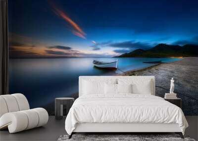 A lone boat sits on a calm, clear water shore at sunset.  A mountain range in the distance is shrouded in mist.  The water is an abstract blur. Wall mural