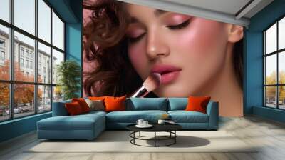 A beautiful woman with long brown curly hair, wearing soft pink eyeshadow and blush on her cheeks is applying powder to the cheekbones of one side of her face using a makeup brush Wall mural