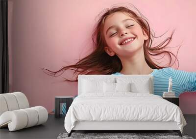 A joyful young girl in a blue striped shirt poses for a studio portrait on a plain pastel pink background embodying a carefree childhood lifestyle with copy space image Wall mural