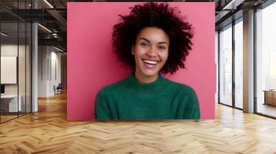 Portrait of good looking curly ethnic woman smiles broadly, enjoys day off, has happy talk with interlocutor, discusses holiday preparation, wears green sweater, isolated on pink background. Wall mural