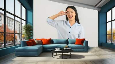 Portrait of Happy young asian woman in blue shirt doing peace symbol with fingers over face isolated on white background Wall mural