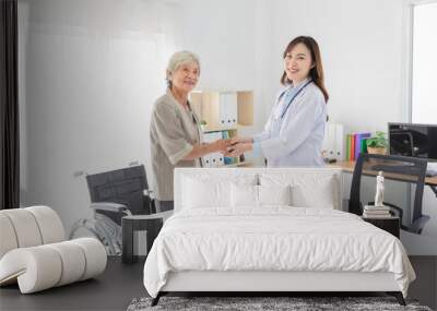 Asian female doctor warmly greeting elderly patient in medical office. They holding hand together, geriatric care, and positive doctor-patient relationship in a clinical setting. Wall mural