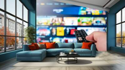 Hand with tv remote aimed towards big 4k flat screen oled tv with blurred out tv entertainment system meny. Technology and quarantine concept. Wall mural