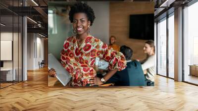 Beautiful young smiling professional black african business woman holding laptop, coworkers hold a meeting in background Wall mural