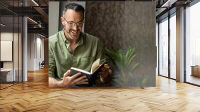 Mature man reading book by window, smiling wearing glasses, copy space Wall mural