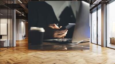 Hands use a mobile phone while working on a laptop in a dimly lit environment Wall mural