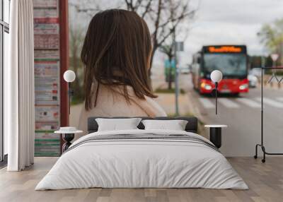 Woman from the back and standing watching her bus arrive at the bus stop. Image of a young brunette girl watching as a red bus approaches her position. Wall mural