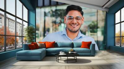 close up portrait of hispanic young entrepreneur smiling and looking at camera at workspace. Front view of one latin happy employee office worker with glasses and successful attitude standing outdoors Wall mural