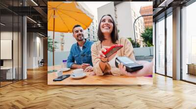 Happy couple paying the bill of the coffee shop using a cellphone contactless payment application. Young adult customer woman doing a purchase on a restaurant with a contactless smartphone banking app Wall mural