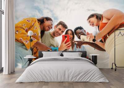 Low angle view of a group of smiling multiracial teenagers addicted to smartphones, watching funny videos, shopping online, enjoying outdoors. Multiethnic cheerful young people searching ,entertaining Wall mural