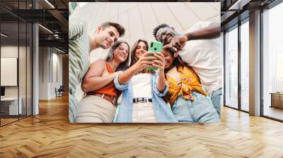 Low angle view of multiracial group of young friends enjoying and smiling using their mobile phone app at university campus. Diverse teenagers having fun watching content on the social media outdoors Wall mural