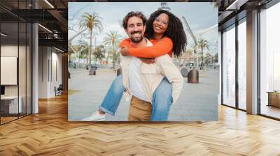 Multiracial couple embracing and having fun. Caucasian man giving his african american girlfriend a romantic piggyback ride enjoying a nice day together. Smiling guy in love carrying on back his woman Wall mural