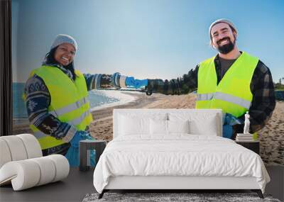 Two multiracial volunteer bumping fists after picking up garbage and plastic dirt from the sand on the beach. A couple of environmental activists smiling standing looking at camera. Ecology concept Wall mural