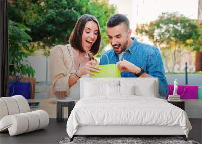 Young adult couple unpacking and looking inside the shopping bags their new purchases. Real people ejoying together at mall. Girlfriend showing to her boyfriend a gift that she buy. Positive customers Wall mural