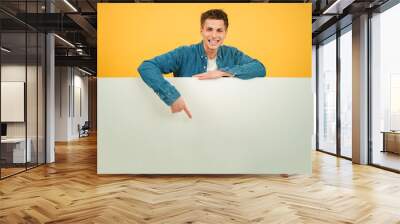 Young caucasian man pointing a blank empty board against red backdrop. Advice copy space mockup. Teenage guy showing a down billboard copyspace at yellow color background wall. Male presenting Wall mural