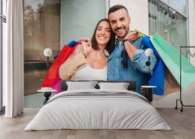 Young couple smiling and looking at camera carrying shopping bags walking on the city mall. Boyfriend and girlfriend laughing near the storefront. Customers buying presents or gifts together Wall mural