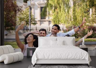 Five happy friends gather for a selfie in a sunny park, smiling, making peace signs, and enjoying a fun moment together, with greenery and urban surroundings.  Wall mural