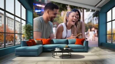 Three friends, two women and a man, are having fun while visiting a city center, laughing at something on a smartphone Wall mural