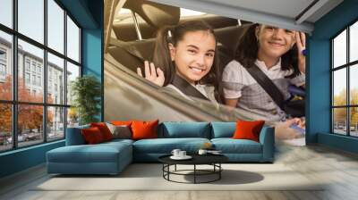 Little girls sitting on a backseat of a car looking camera going to school by car and waving goodbye. Transportation concept routine morning. Wall mural