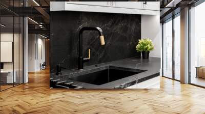 A kitchen sink detail with a modern black and gold faucet, with a black stone countertop and backsplash, and white cabinets. Wall mural