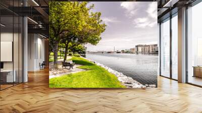 Beautiful landscape scenery shot along the Fox River just after sunrise in Green Bay Wiscosin with riverwalk trail, bench, trees, grass and rocky shoreline framing the shot and blue cloud filled sky. Wall mural