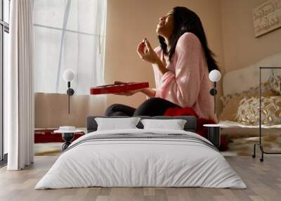 happy african american woman eating romantic valentines chocolates from heart shaped box while sitting on bed at home Wall mural