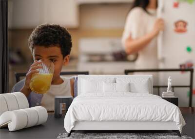 little boy drinking orange juice at breakfast with mother in background Wall mural