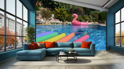 Swimming pool with young woman floating on an inflatable pink flamingo, checking her phone, and  bright rainbow coloured pool noodles in the foreground on a sunny summer day Wall mural