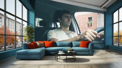 Young man driving car on urban city. Smiling driver holding steering wheel with no seat belt on. Road trip, auto rental, insurance, traffic ticket, irresponsible concepts Wall mural