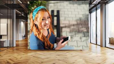 a young girl with orange hair in the park sits on a bench and looks at her mobile phone Wall mural